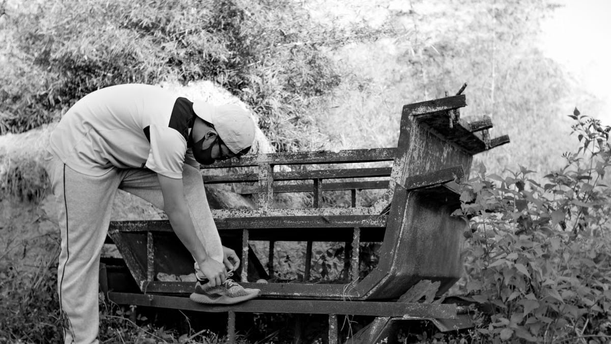 Black and white of male in sportwear and mask lacing up sneaker putting leg on wooden bench near trees and bushes in park